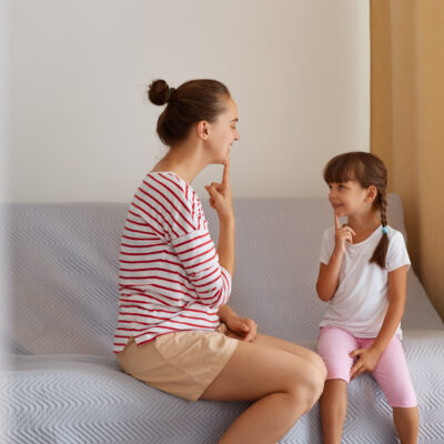Indoor shot of woman with hair ban sitting on sofa with little girl, demonstrating for child how to pronounce sounds, private lesson with professional speech pathologist.