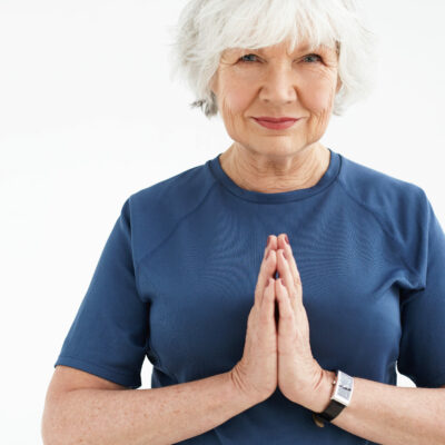 Positive energetic elderly woman with gray hair choosing active healthy lifestyle, smling, holding hands in namaste while practicing yoga or meditation against blank copyspace studio wall background